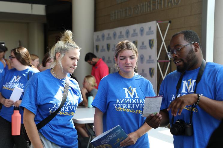 Three students wearing Madonna University t-shirts at orientation are listening to each other speak and looking at a piece of paper intently