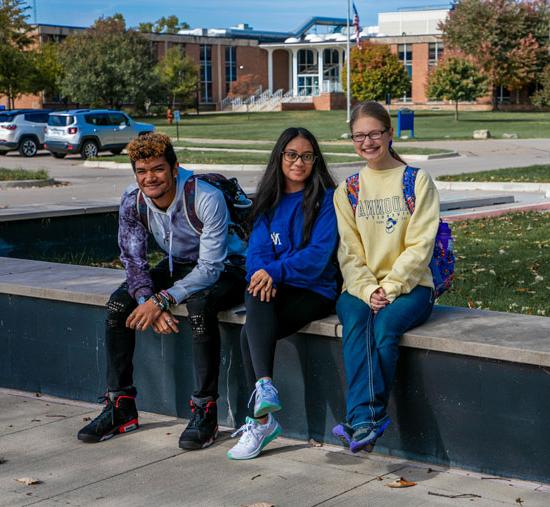 Three smiling students sitting along the short brick wall outside the Welcome Center